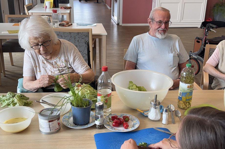 Mehrere Bewohnerinnen und Bewohner bereiten gemeinsam einen Salat zu. Auf dem Tisch liegen Salat, Kräuter, Gemüse und Zutaten für das Dressing.