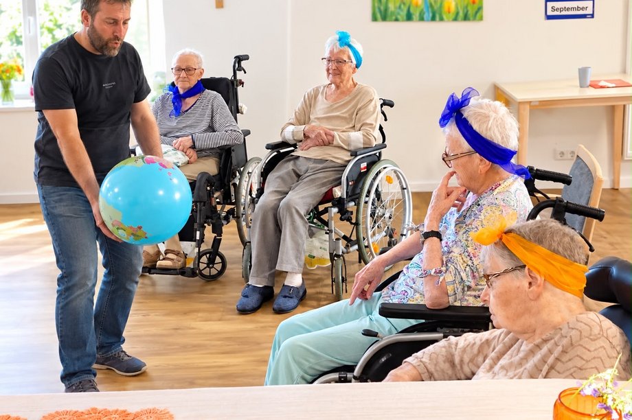 Ein Mann hält einen großen Ball in seinen Händen, um ihn herum vier Seniorinnen im Rollstuhl.