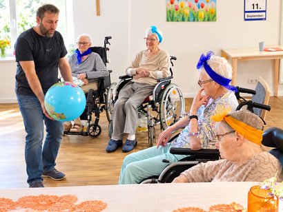 Ein Mann hält einen großen Ball in seinen Händen, um ihn herum vier Seniorinnen im Rollstuhl.