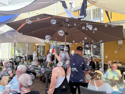 Seifenblasen schweben über einer Terrasse, auf der Seniorinnen und Senioren gemeinsam ein Sommerfest unter großen Sonnenschirmen feiern.