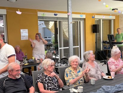 Seniorinnen und Senioren sitzen bei einem Sommerfest auf einer Terrasse. Im Hintergrund spielt ein Musiker Keyboard, einige Gäste klatschen mit.