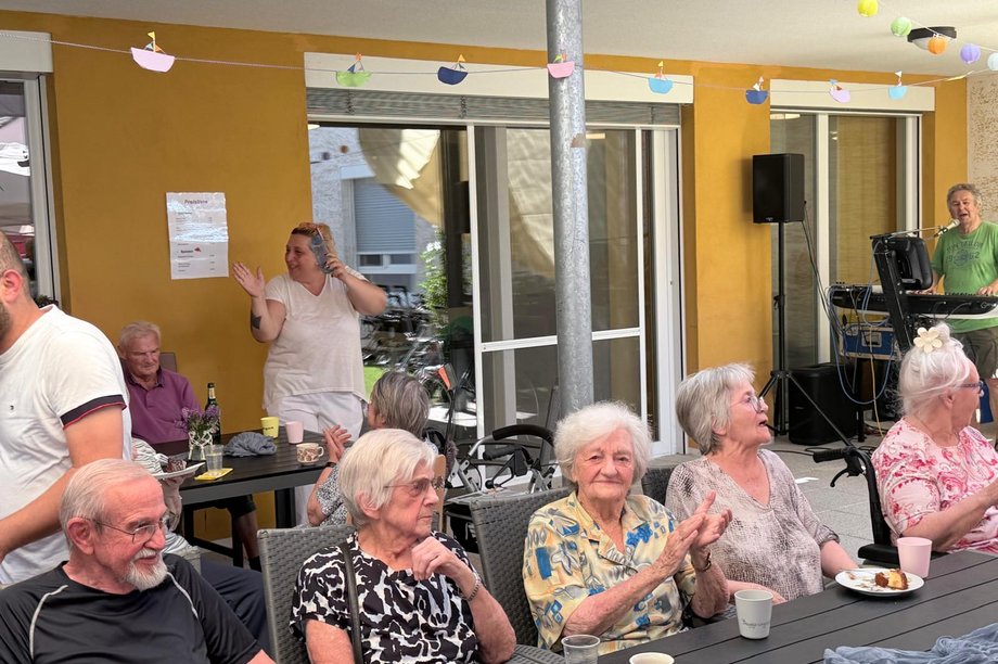 Seniorinnen und Senioren sitzen bei einem Sommerfest auf einer Terrasse. Im Hintergrund spielt ein Musiker Keyboard, einige Gäste klatschen mit.